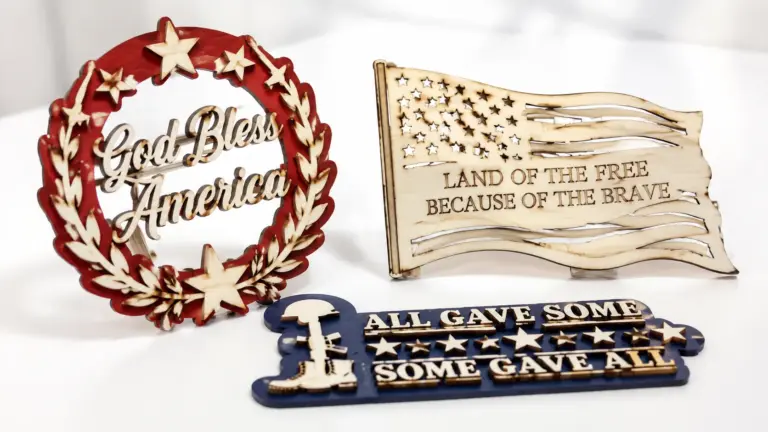 Laser-cut patriotic wood signs made from 3mm basswood, displayed on a workshop table with red, white, and blue finishes